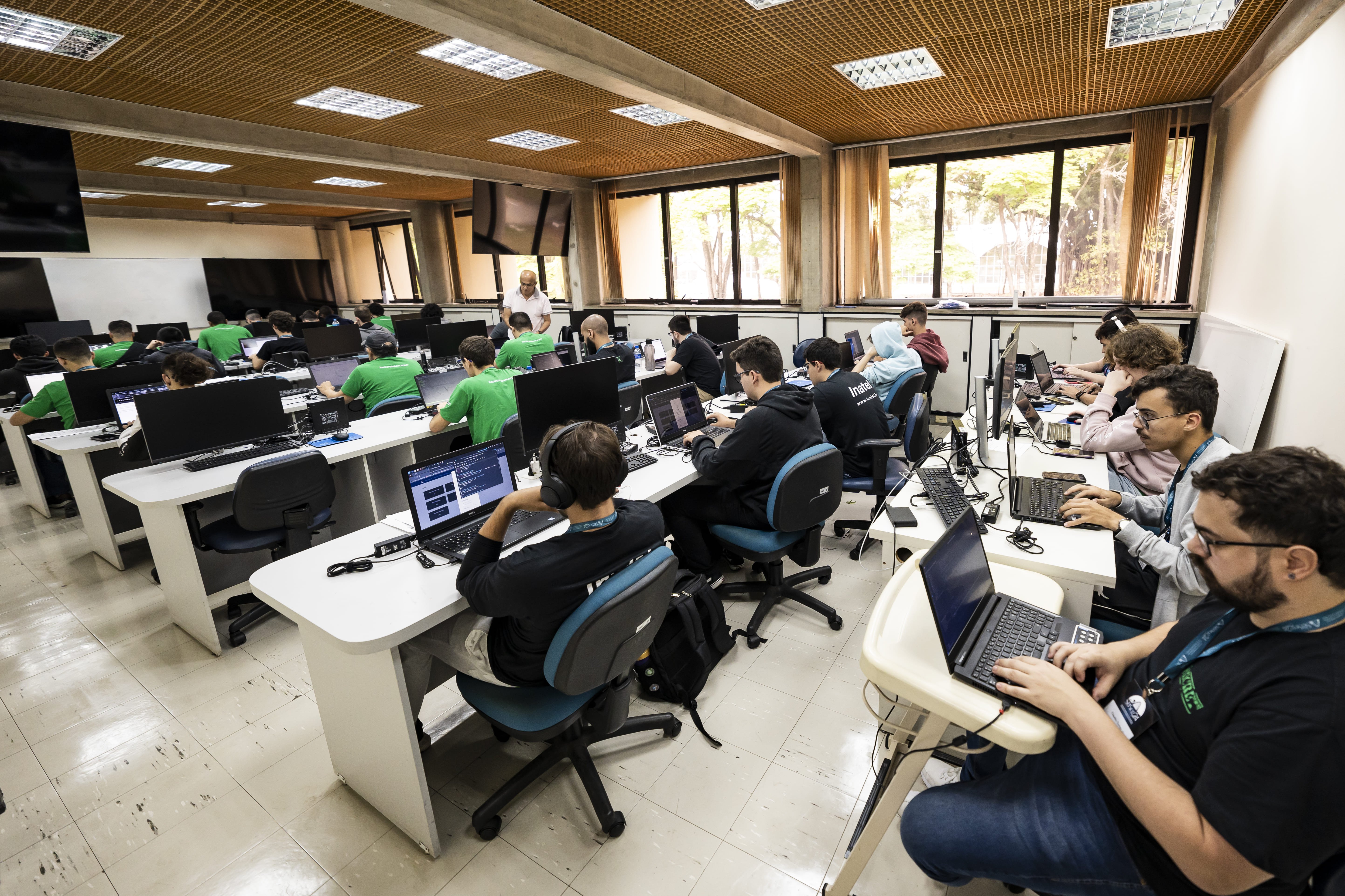 Muitos meninos e uma menina em uma sala cheia de computadores, eles estão concentrados.